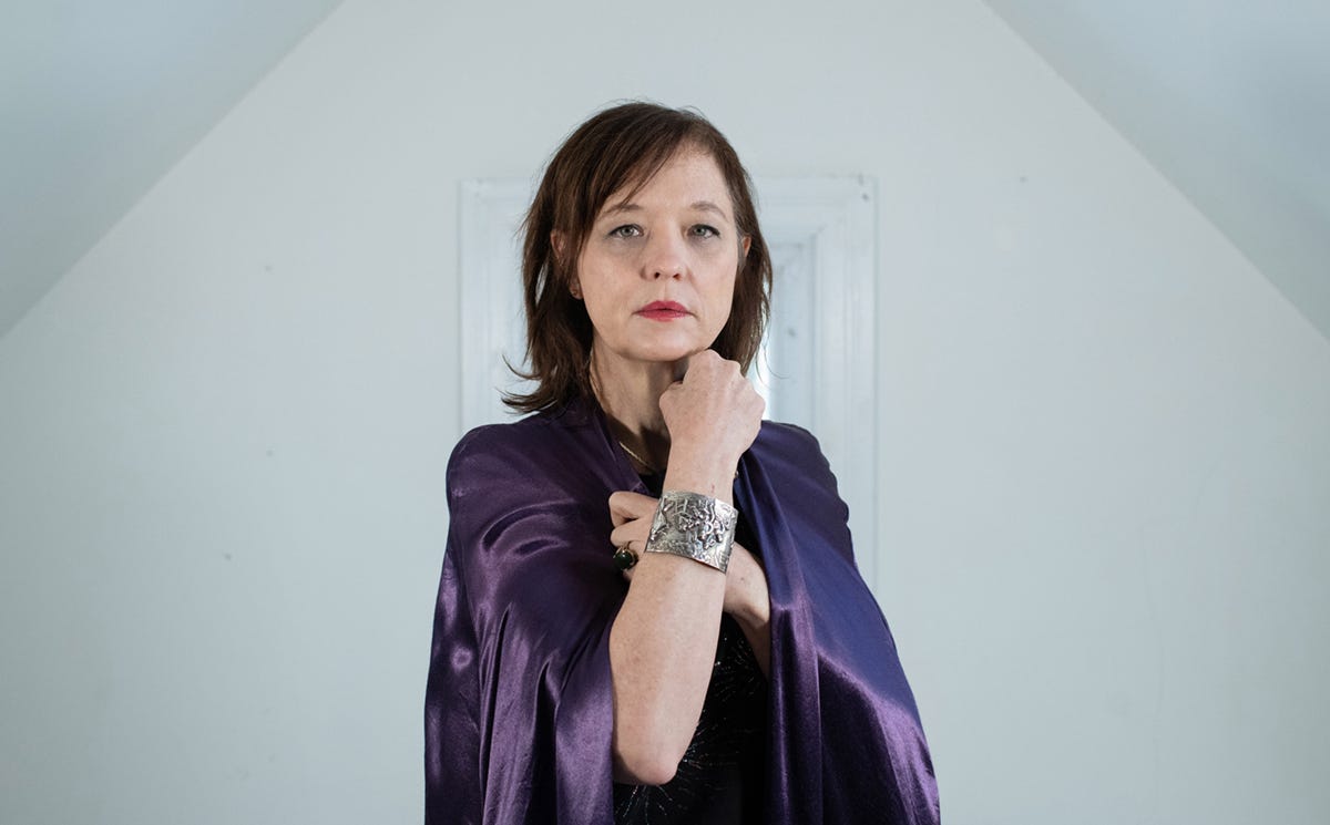 Mary Timony in a purple cape with raised fist showing an elaborate silver bracelet