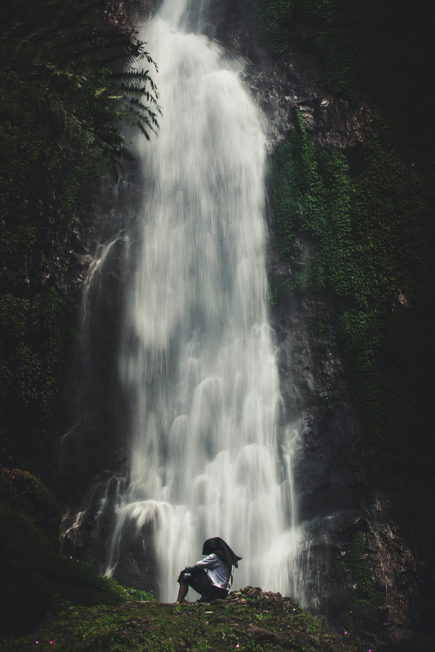 Person sitting at the foot of a large waterfall in a rainforest