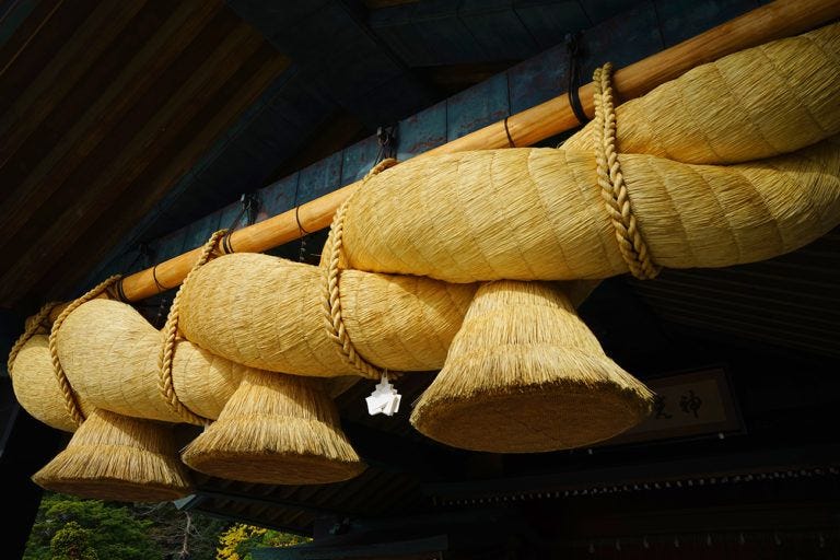 Sogas de paja de arroz (shimenawa) colgadas en un santuario sintoísta, con un diseño trenzado y extremos en forma de campana. Se aprecia su color dorado y la textura de la paja, simbolizando la conexión con lo divino y la tradición japonesa.