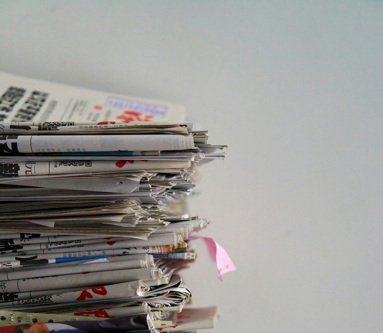 a stack of many newspapers, lying flat on top of each other, against a white wall background