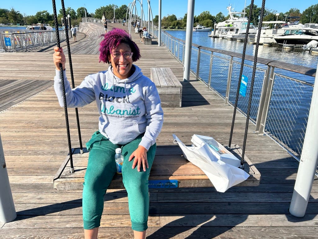Kristen is facing the camera,  sitting on a large swing on a large dock. She's wearing an I Am a Black Urbanist sweatshirt