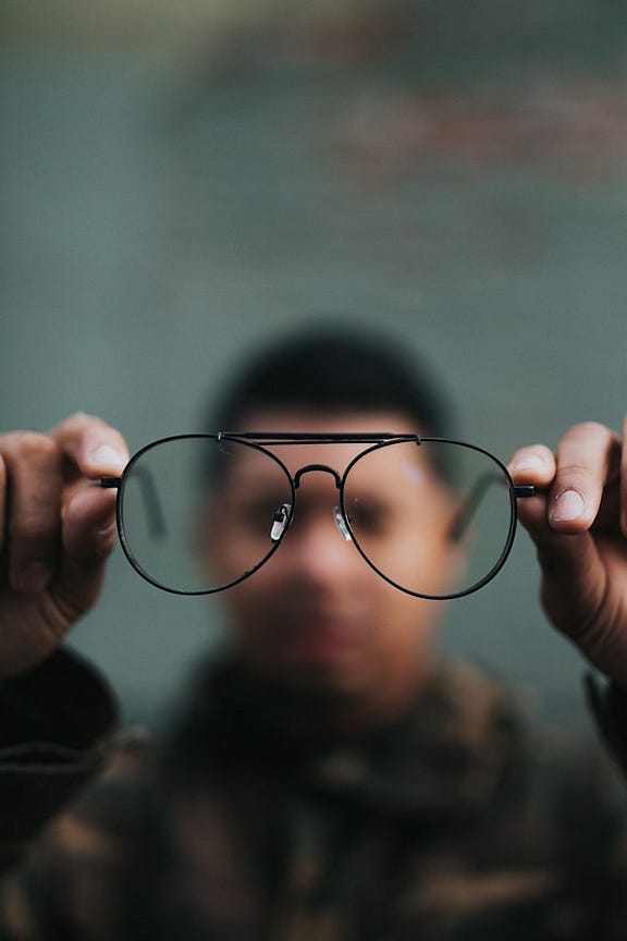 a man holding up a pair of eyeglasses while appearing blurry in the background. The eyeglasses appear to have no lenses, as the man is still blurry in the background