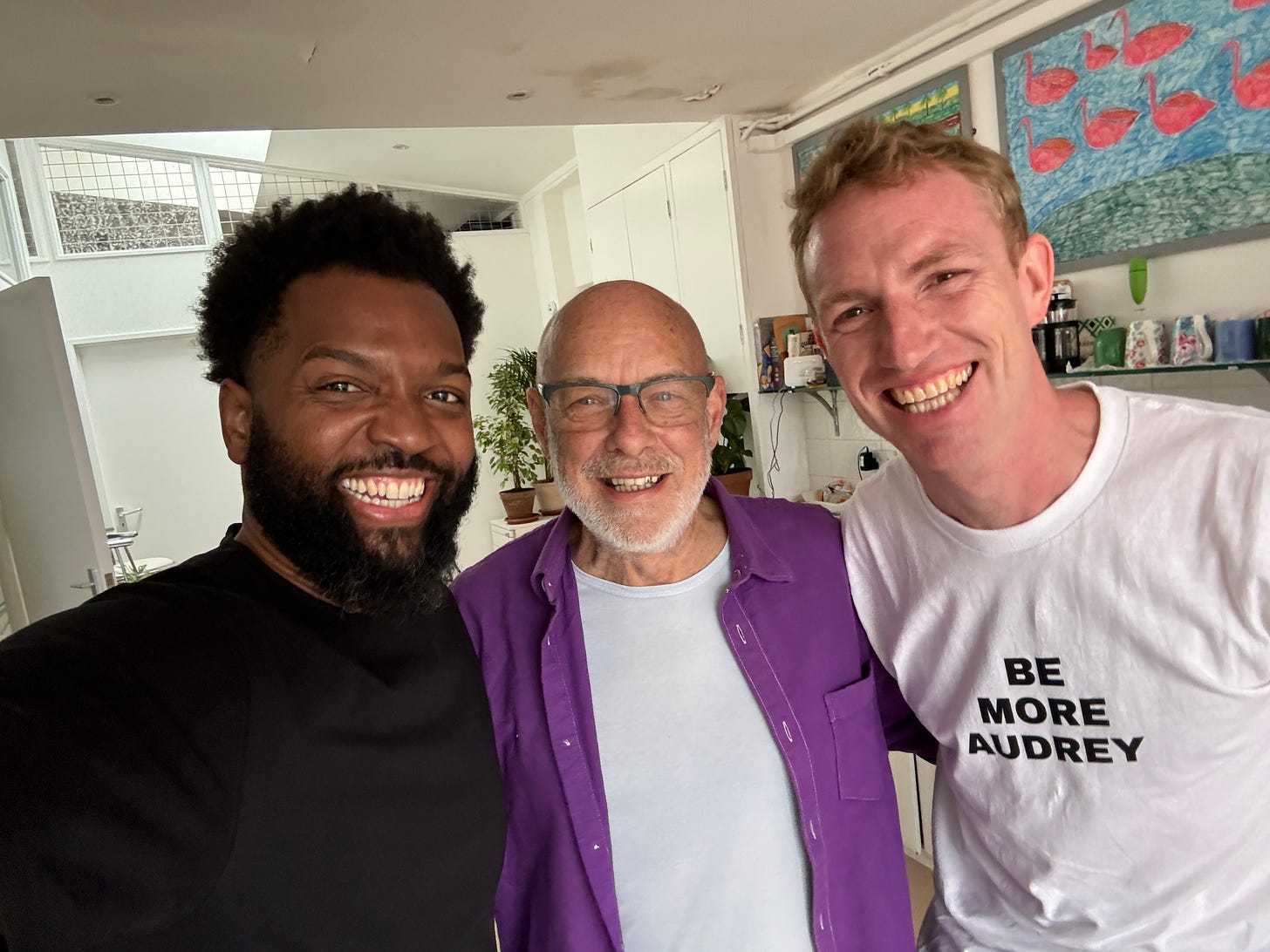 Three smiling men pose together indoors. The man on the left (Baratunde) has a beard and wears a black shirt. The man in the middle (Brian Eno) is older, bald with a white beard, and wears glasses, a light blue shirt, and a purple overshirt. The man on the right (Jon Alexander) has short blond hair and wears a white T-shirt with the text “BE MORE AUDREY.” Colorful artwork and shelves are visible in the background.
