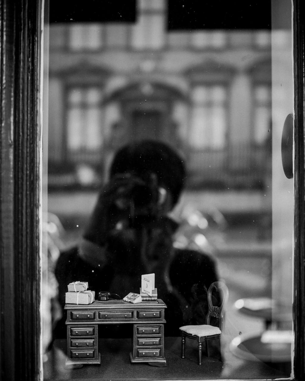 Miniature desk, chair and books in a shop window, with the photographer and building across the road reflected in the window, in black and white