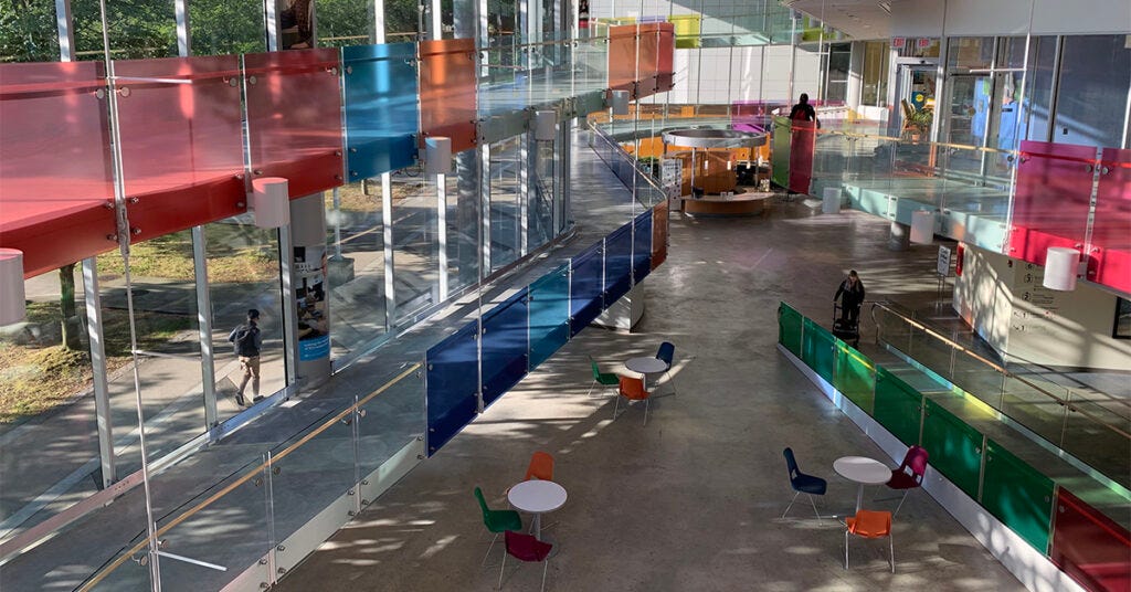 an aerial shot of the lobby in the Blusson Centre, with long sloped ramps between floors and colorful semi-transparent glass lining the rampways. Light fills the room from an all-window wall on one side.