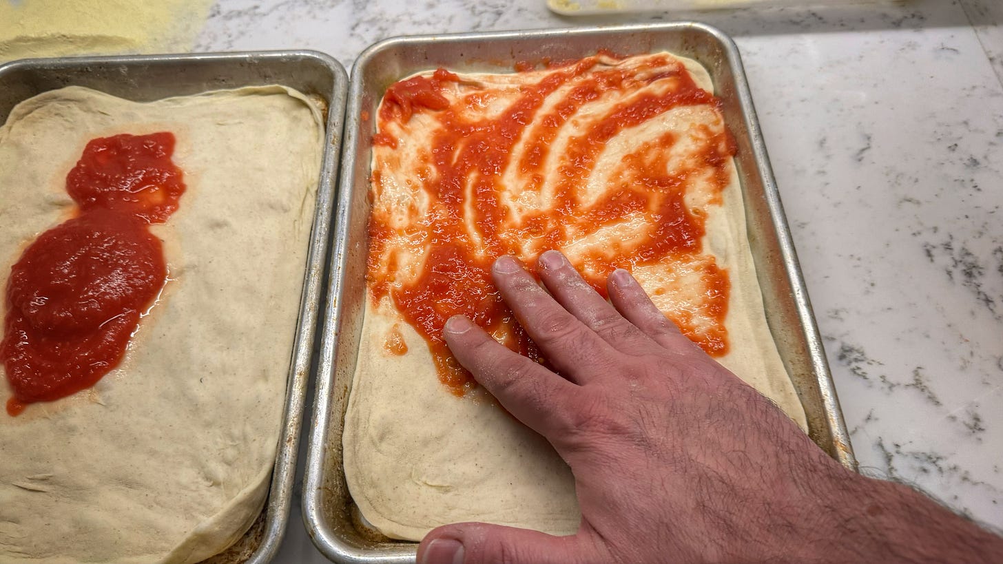 A hand smearing tomato sauce onto a pizza dough stretched to fit a quarter sheet pan.