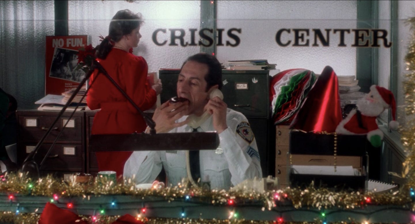 A screengrab from Home Alone. A man sits at a desk talking on a chorded phone while he takes a big bite out of a donut. The office is decorated for Christmas. A woman in a red dress potters around in the background.