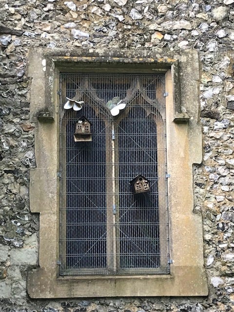 Stony East Wall of the Chaldon Church, highlighting a window, protected by a grille. There are bird feeders and butterflies made of what might be metal, attached to the window grille