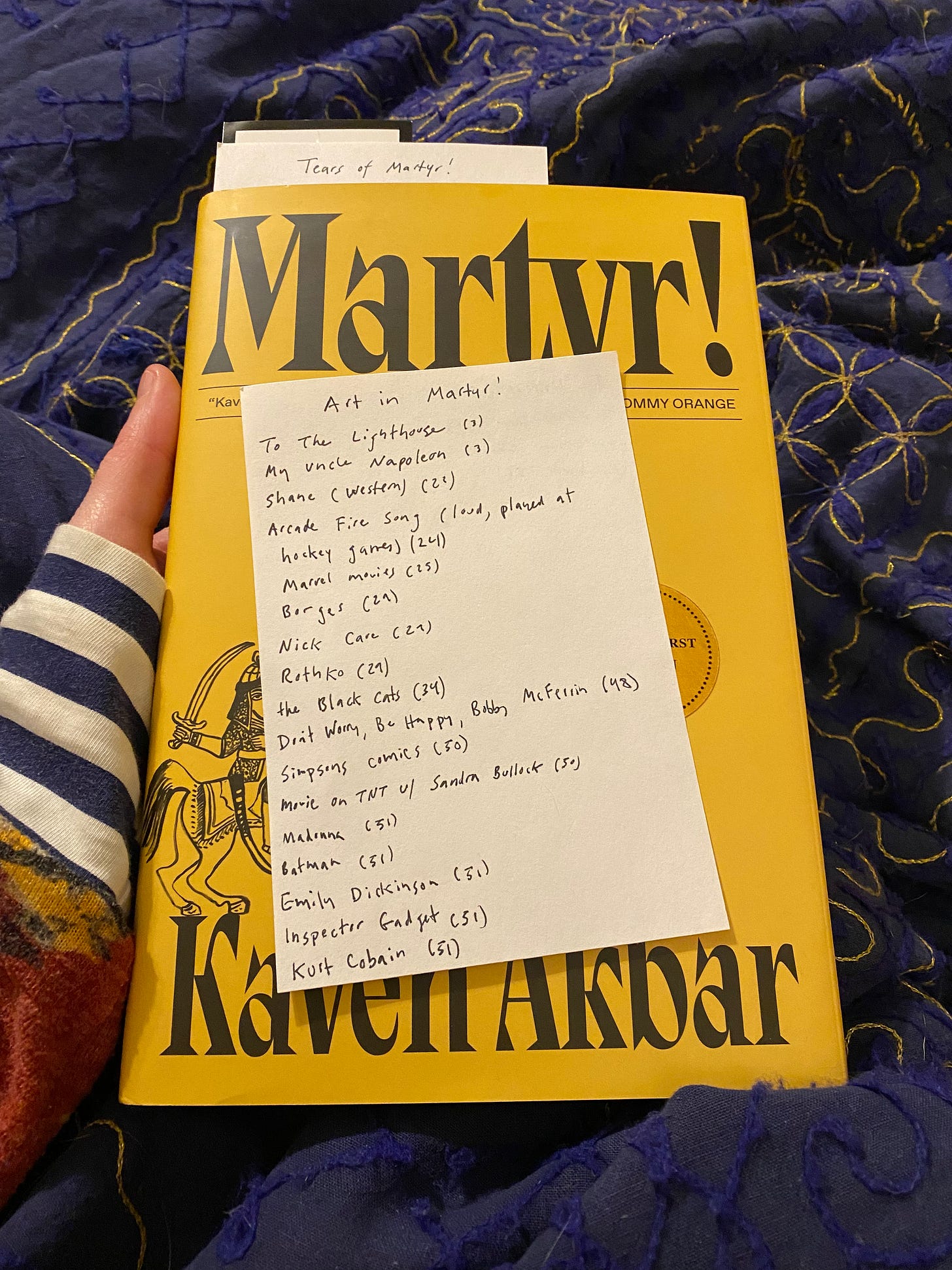 I’m holding the yellow hardcover of Martyr! An index card with a list of books, films, songs, artists, etc., along with page numbers, rests on top of the book.