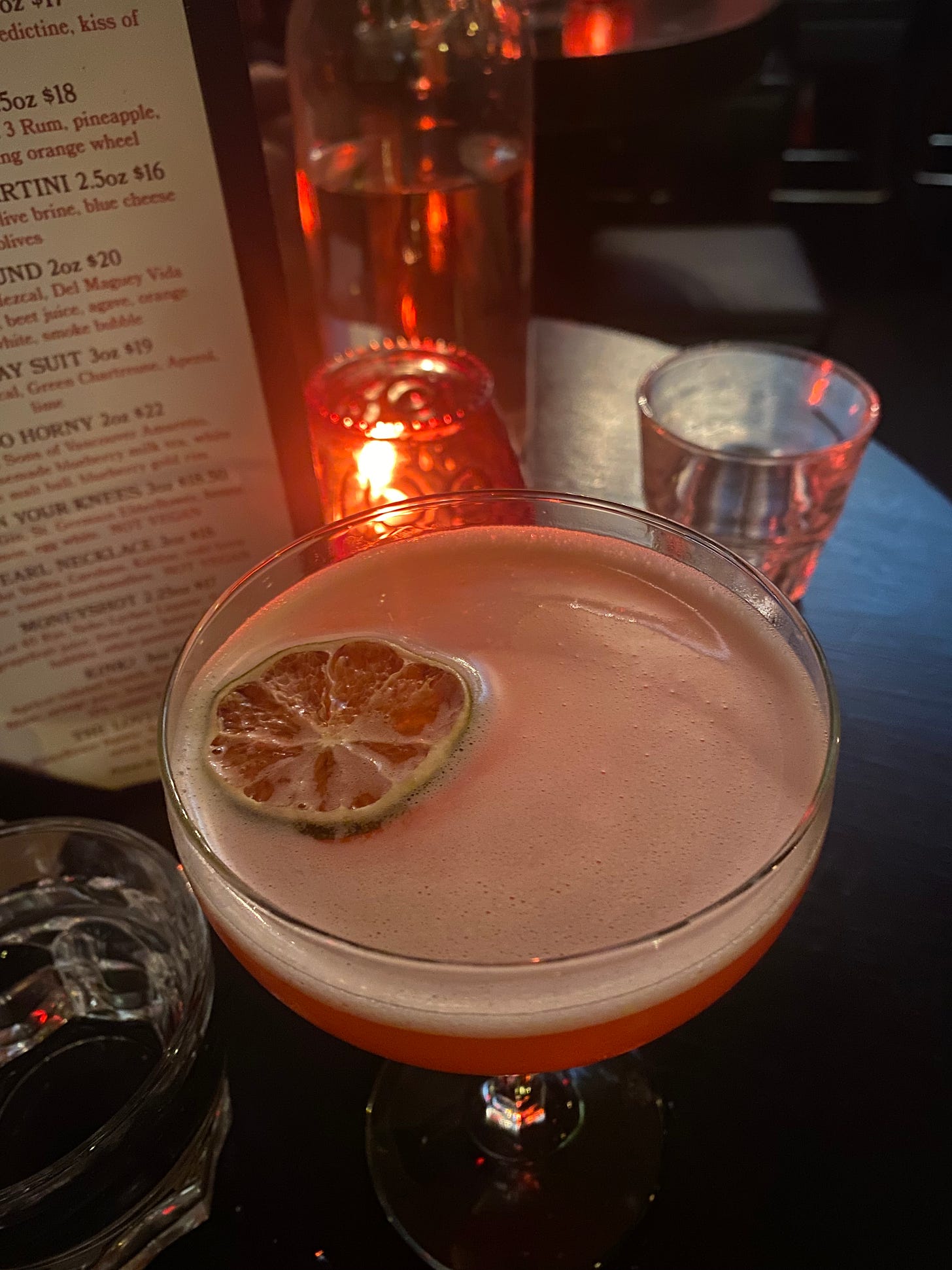 A pink-ish cocktail in a coupé glass with white foam and a dehydrated lime slice. In the background is a candle in a red textured class, and two small glasses of water.