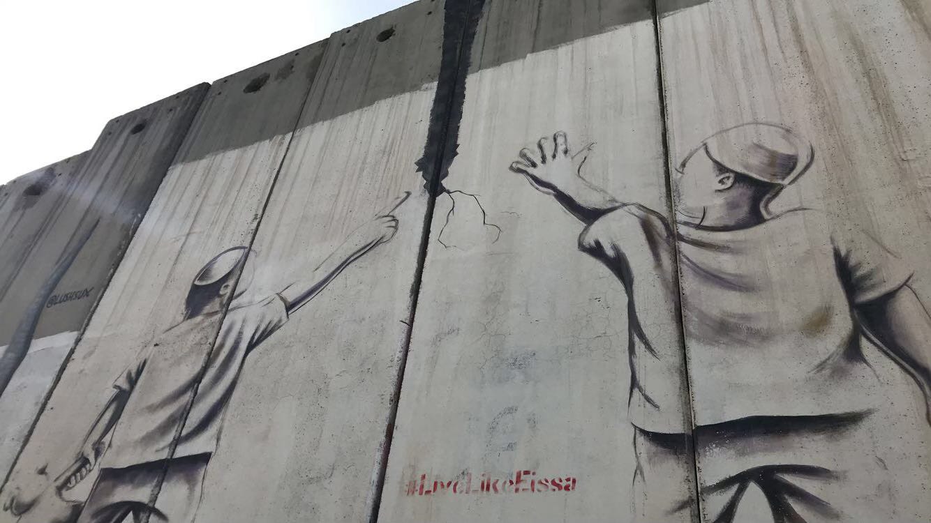 Graffiti of two boys pointing to a crack in the separation wall in Bethlehem, Palestine. Photo by Riad Alarian, 2018.