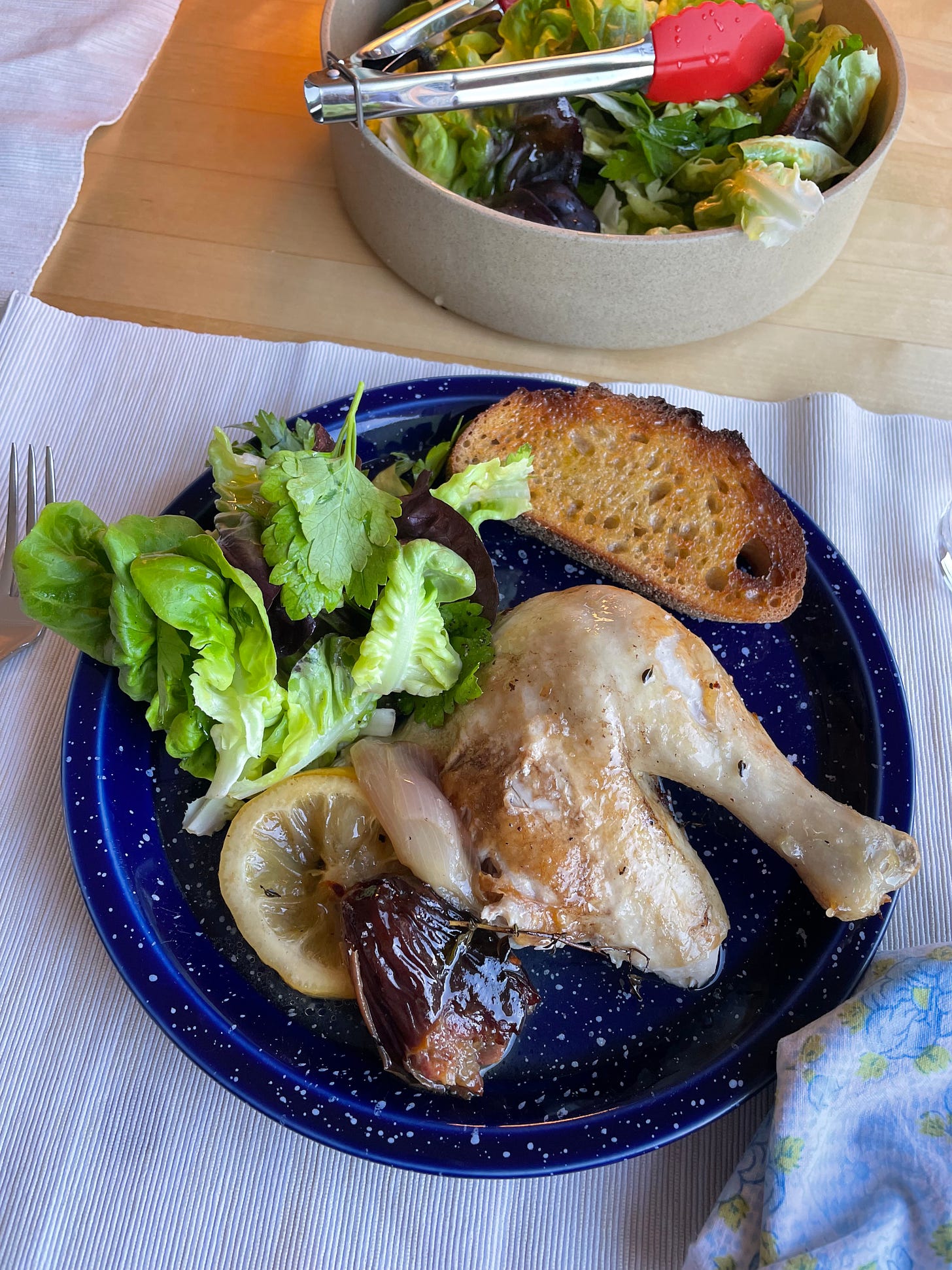 A speckled plate with a green herby salad, and a chicken thigh and drumstick, with a sticky date and buttered toast sitting alongside it. A ceramic bowl of green salad in the background.