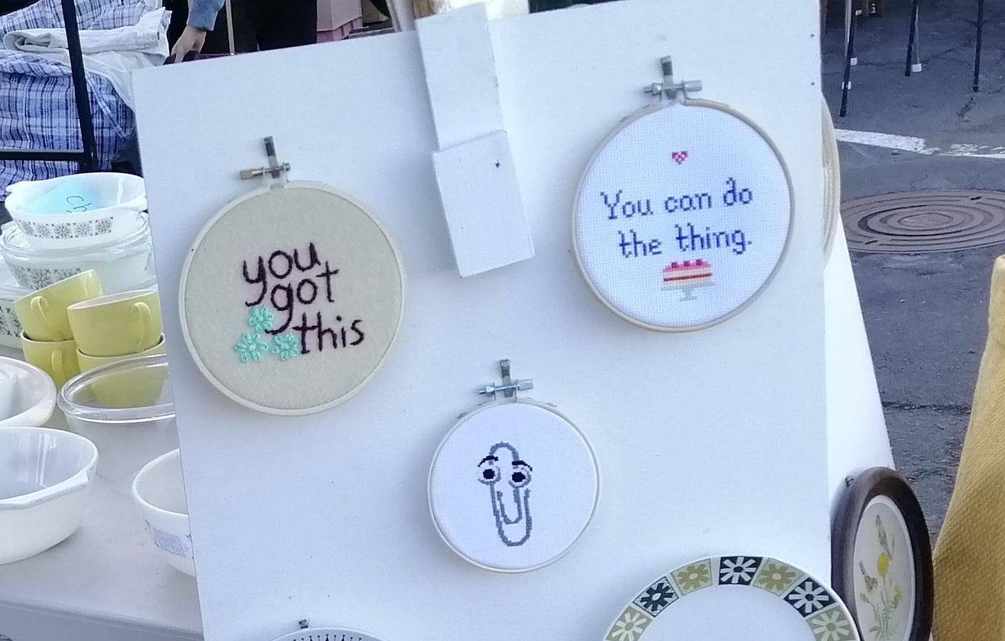 Cross stitches in hoops on a market stand, one reads "you got this", one "you can do the thing" with a picture of a cake, and one of Clippy, the Microsoft Word Assistant from ye olden times