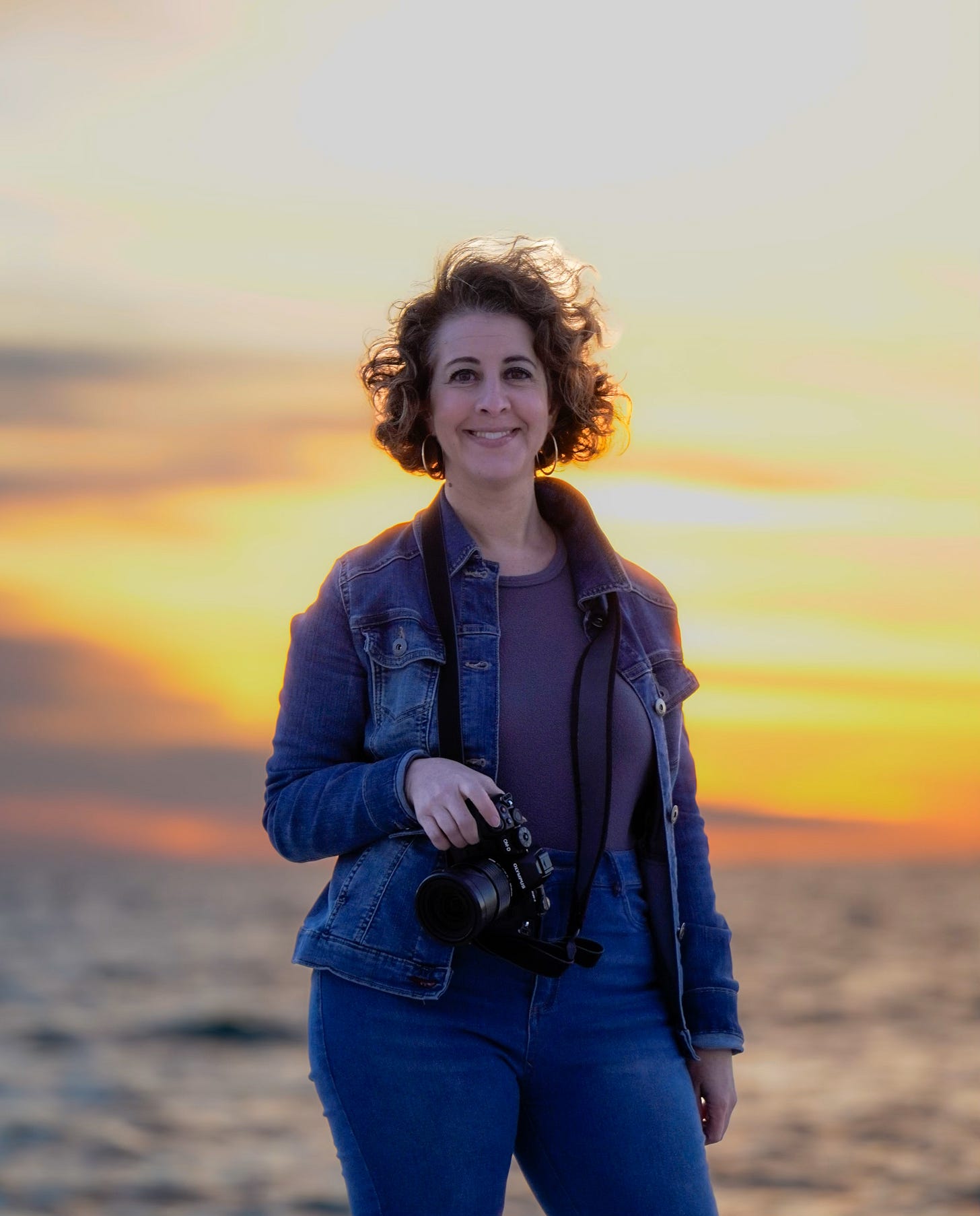 Woman with short brown curly hair holds her Nikon camera while the sunsets in the background