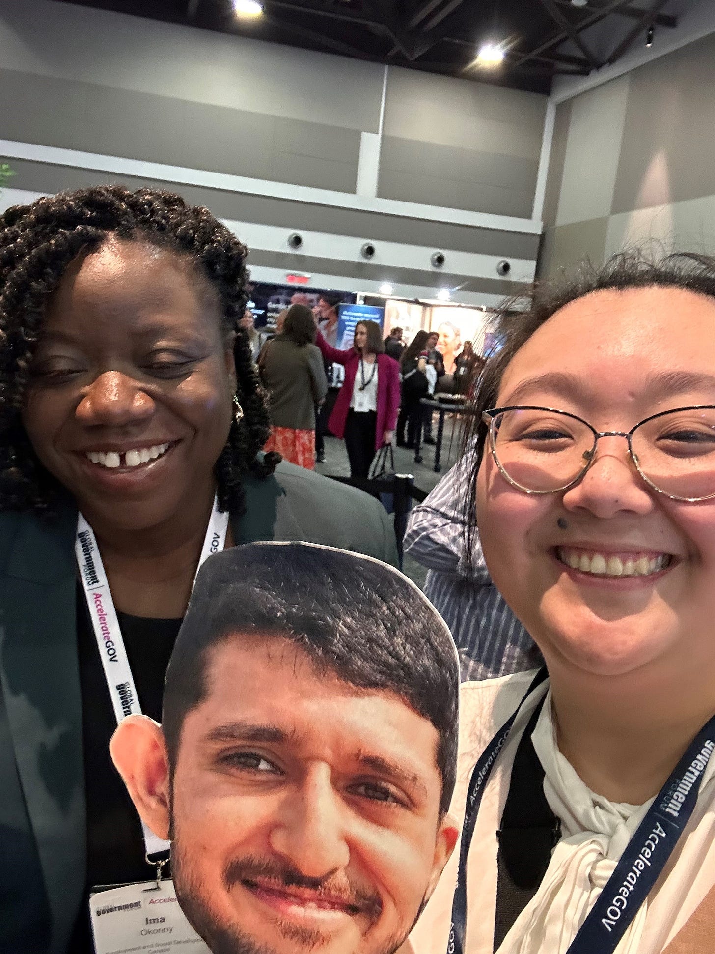 Heidi and Ima Okonny smile widely at the camera. In the background is the kiosks of AccelerateGov with participants talking. Heidi is holding a cardboard cutout of Lajendon.