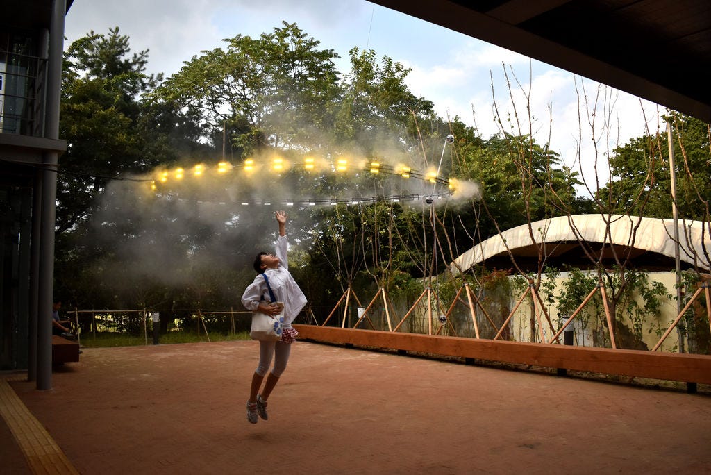 Photo of an East Asian man in a courtyard is mid jump, reaching into the air where a giant illuminated halo with yellow lights emits water vapour into the air.