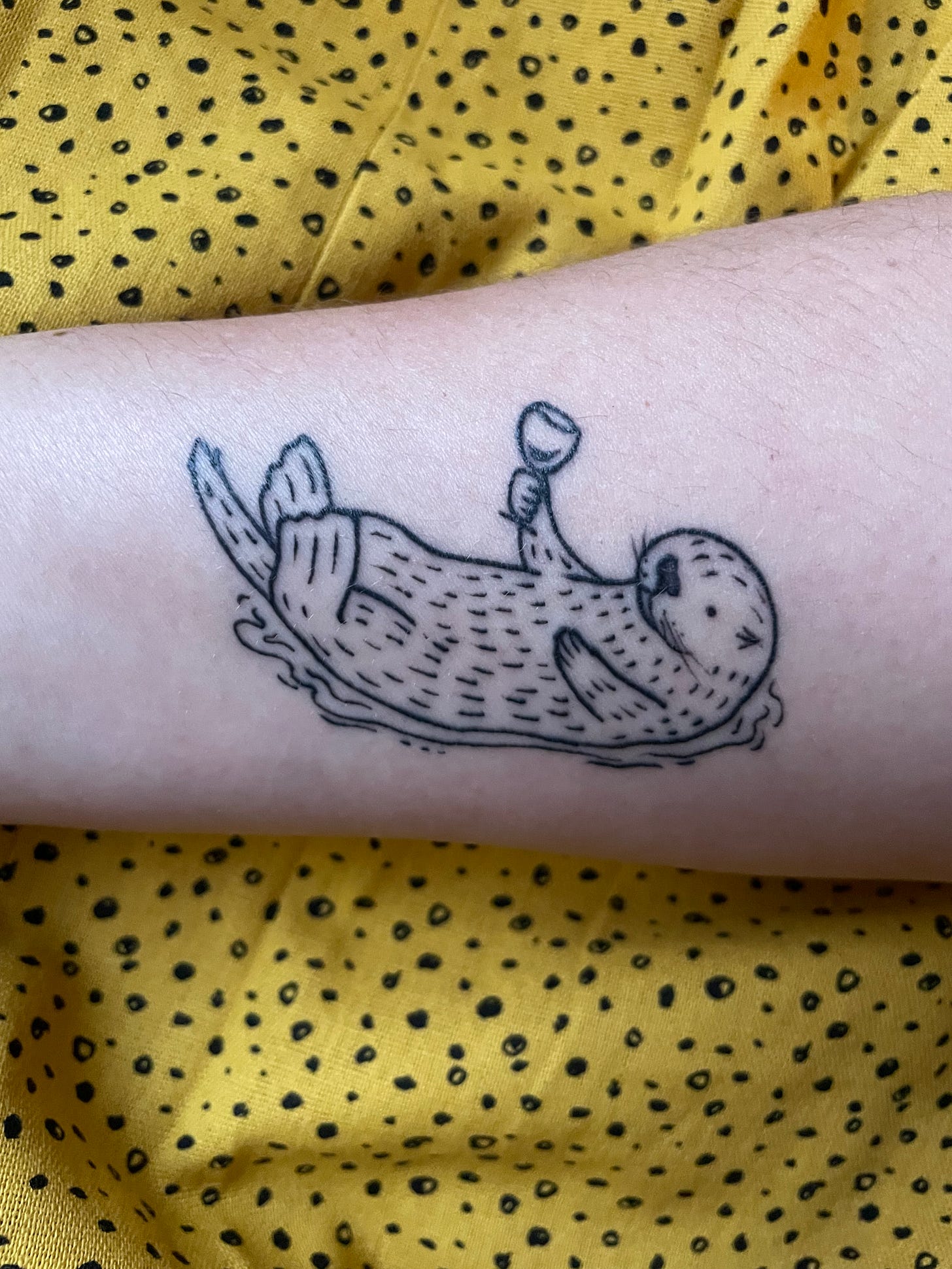 A tattoo of a sea otter drinking a glass of wine on olive-toned skin. 