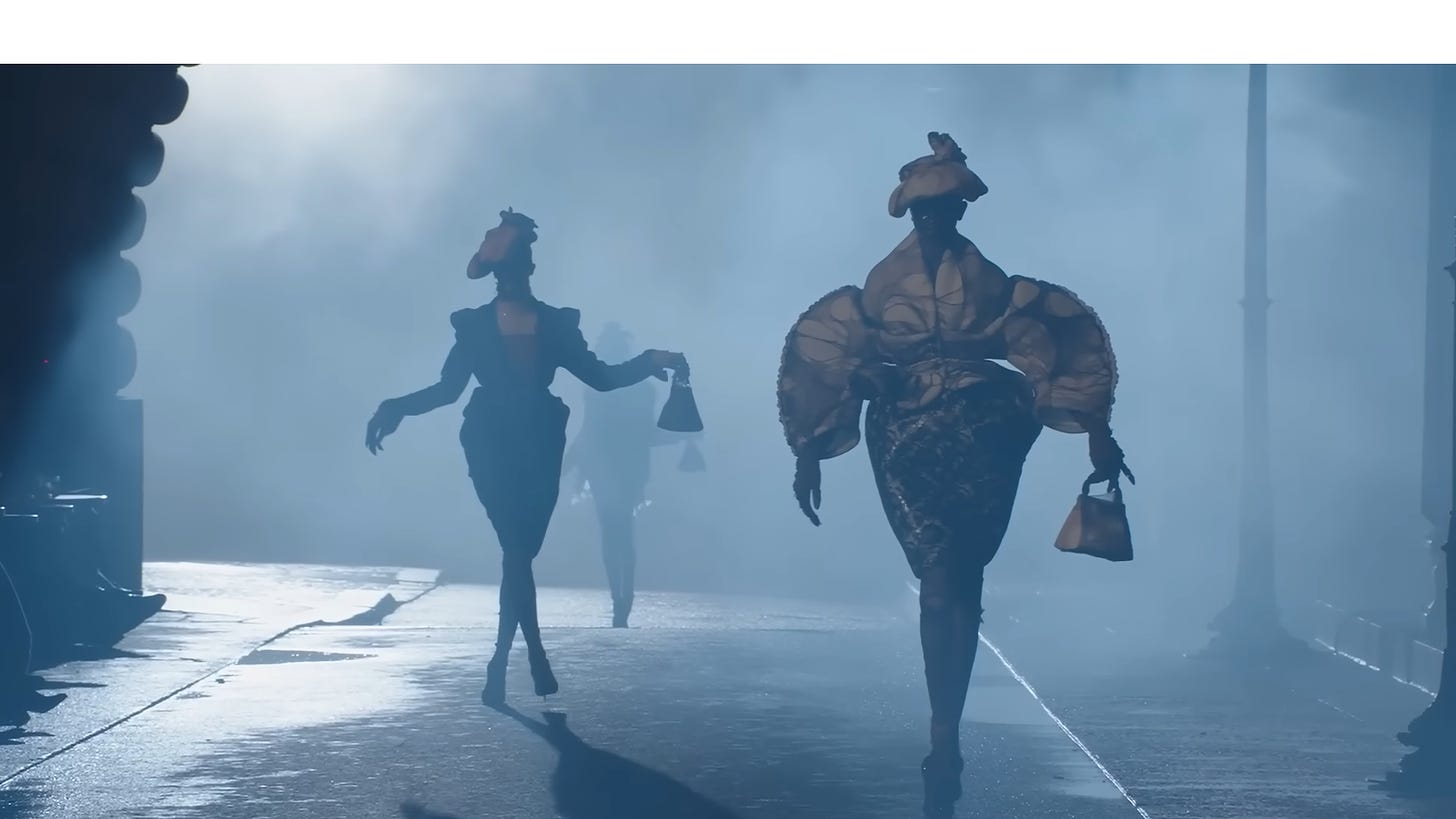 Immagine della sfilata Margiela. In un vicolo nebbioso, le silhouette di due modelle (una in primo piano, l'altra in secondo piano) camminano con fare teatrale. 
