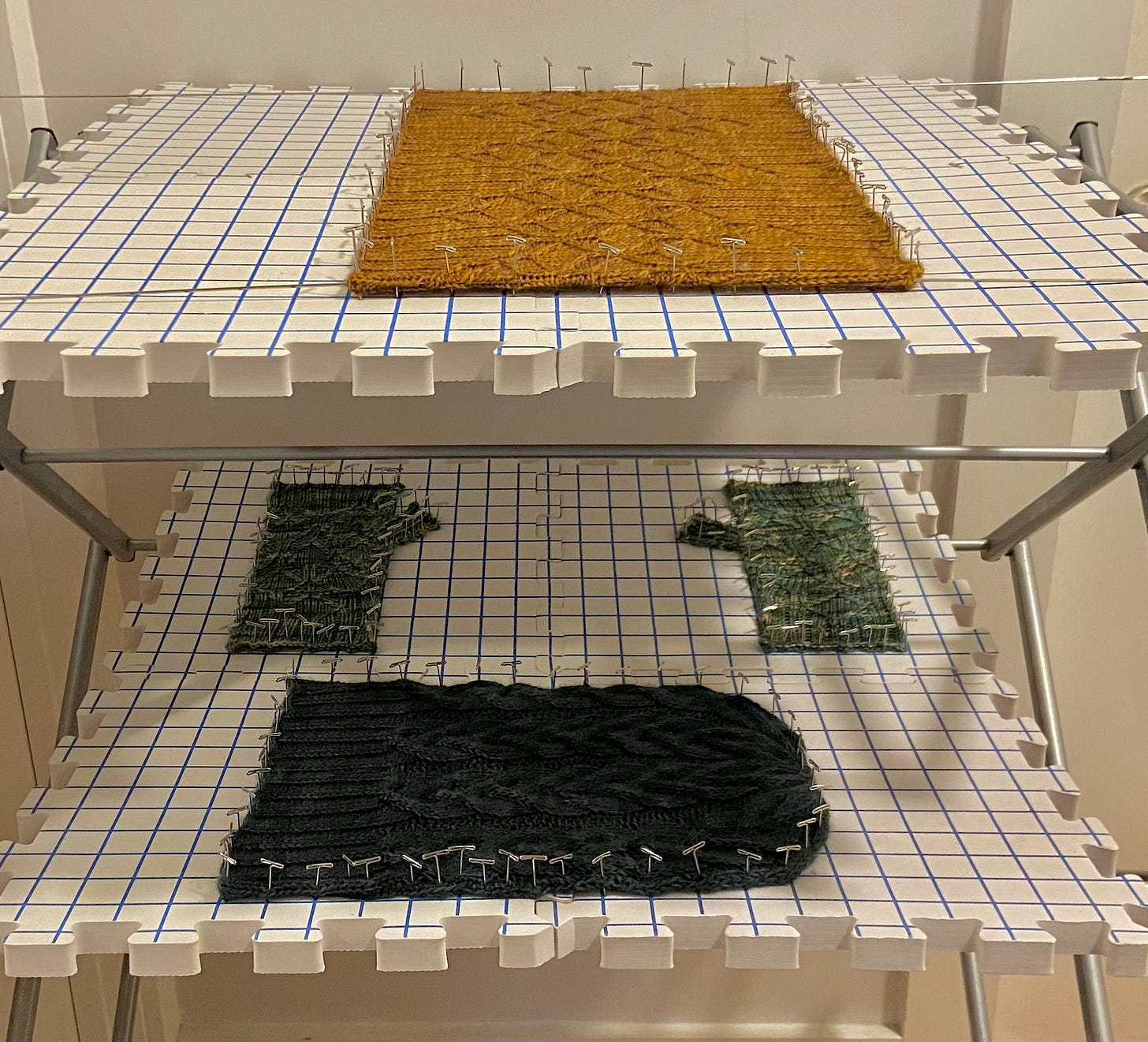 (top to bottom) An orange cowl, greenish-blue fingerless gloves, and a green hat all pinned and blocked on two foam-like puzzle pieces used for blocking knitted garments. They're resting on two rows of a drying rack. The cowl has two metal bars to help with the blocking.