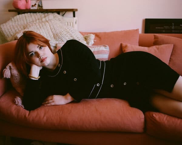 Eliza Clark, wearing a black dress, reclines on a pink sofa and looks directly at the camera.