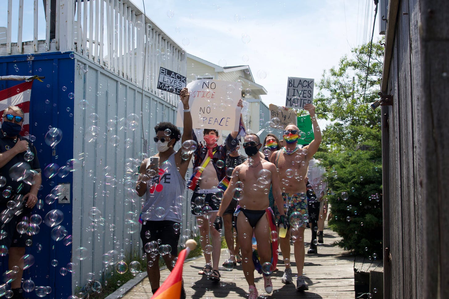 A group of diverse protesters on a wooden boardwalk, holding "Black Lives Matter" and "No Justice No Peace" signs. Many of the protesters wear colorful clothing, sunglasses, and protective face masks. A myriad of shimmering soap bubbles float around them, creating a whimsical atmosphere against the backdrop of a white and blue fence and a clear sky.