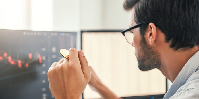 A photo of a man looking at cryptocurrency price data on a computer screen with a coin between his fingers.