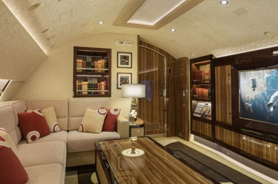 Interior of a Boeing 747 private jet, showing a seating area with a large screen TV and bookshelves.