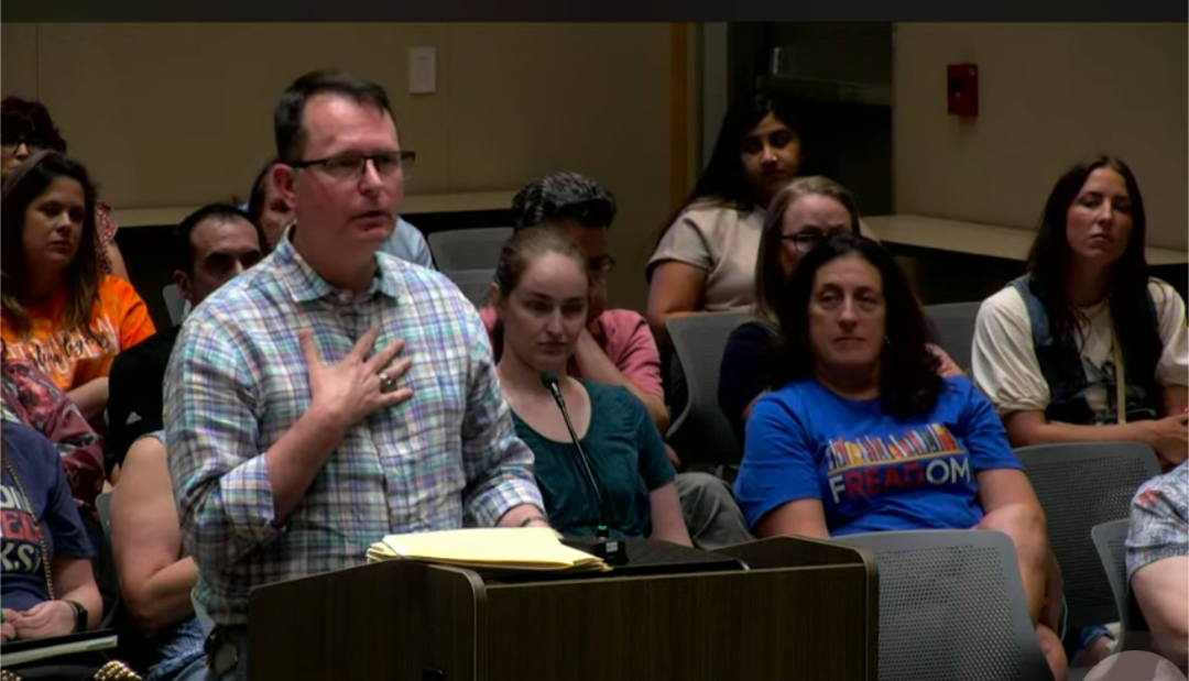 Parent standing at podium in front of seated audience at school board meeting, clutching at his neck as if he's grasping his pearls.