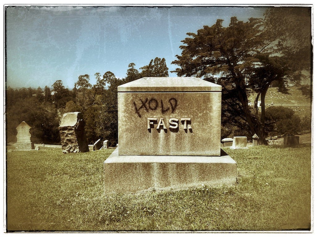 A vintage photograph of a tombstone in a graveyard. The name on the tombstone reads, "Fast," and someone has spray-painted the word, "Hold," above it."