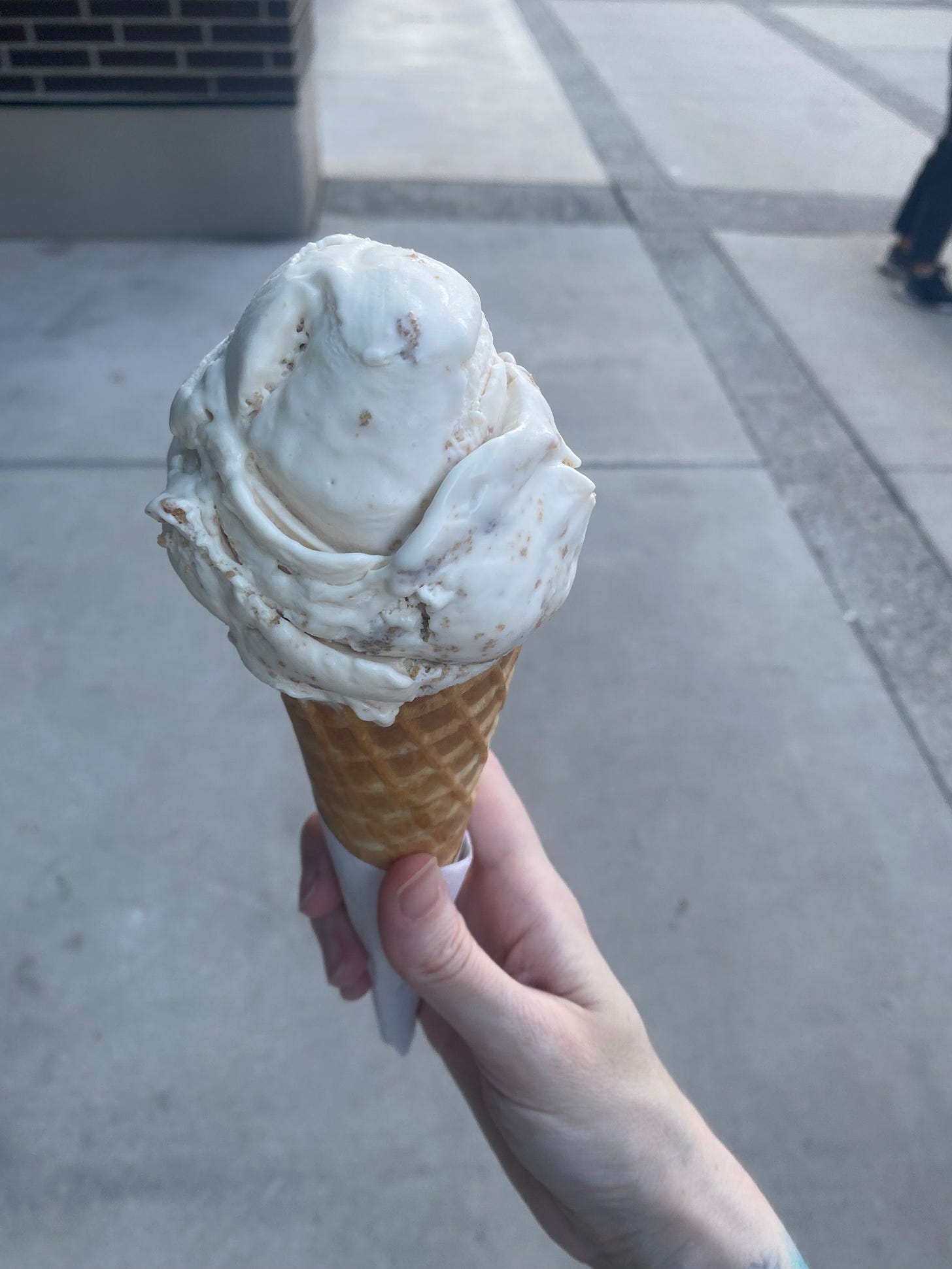 My hand, holding a waffle cone of the key lime pie ice cream described above. It's white with graham crumbs visible throughout.