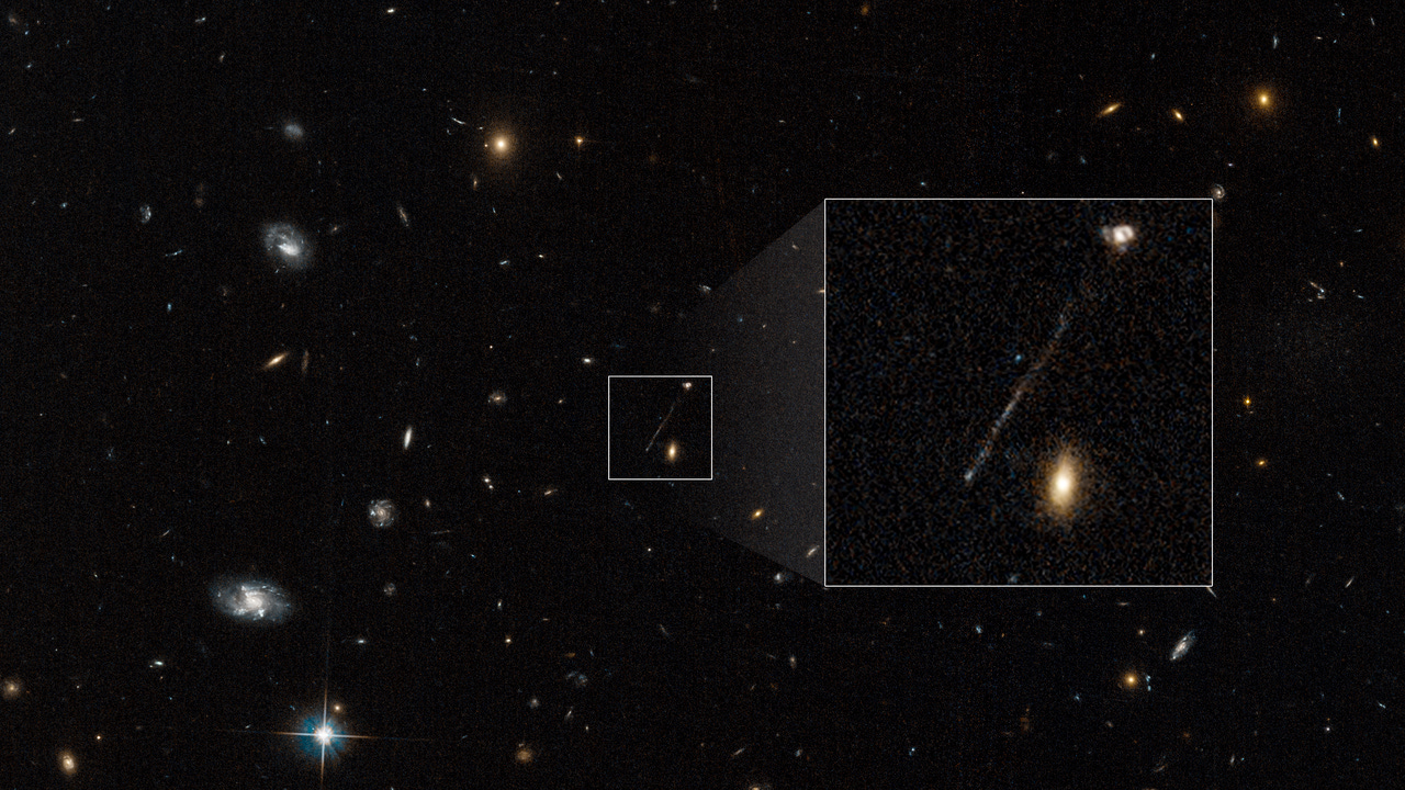 A Hubble image of a black, deep-space field is speckled with white, yellow and reddish galaxies. At the lower left corner, one, lone star with four, prominent spikes appears. In the center of the image is a small, white-bordered, boxed area that contains one, long, thin, diagonal streak of whitish-blue stars. The diagonal line looks like a scratch on the photograph. It is positioned from near the bottom, left corner to the top right corner of the box. Two galaxies also reside within the box. One irregular-looking galaxy, in the top right corner, appears to be connected to the line. Another elliptical-looking galaxy appears in the bottom, right corner of the box. To the right of the small box is a larger white-bordered box that contains a magnified view of the contents of the smaller box. In this magnified view, a white knot appears prominently at the bottom tip of the stream of stars.