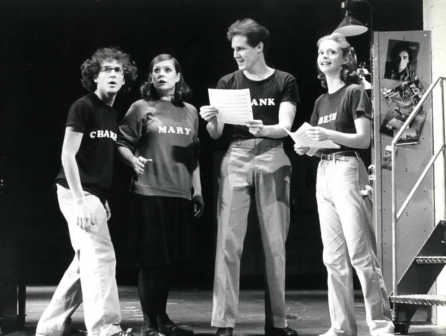 Black and white photograph of the cast in a scene from Merrily We Roll Along. The main characters are wearing sweatshirts with their names on them, standing next to a set resembling a high school locker.