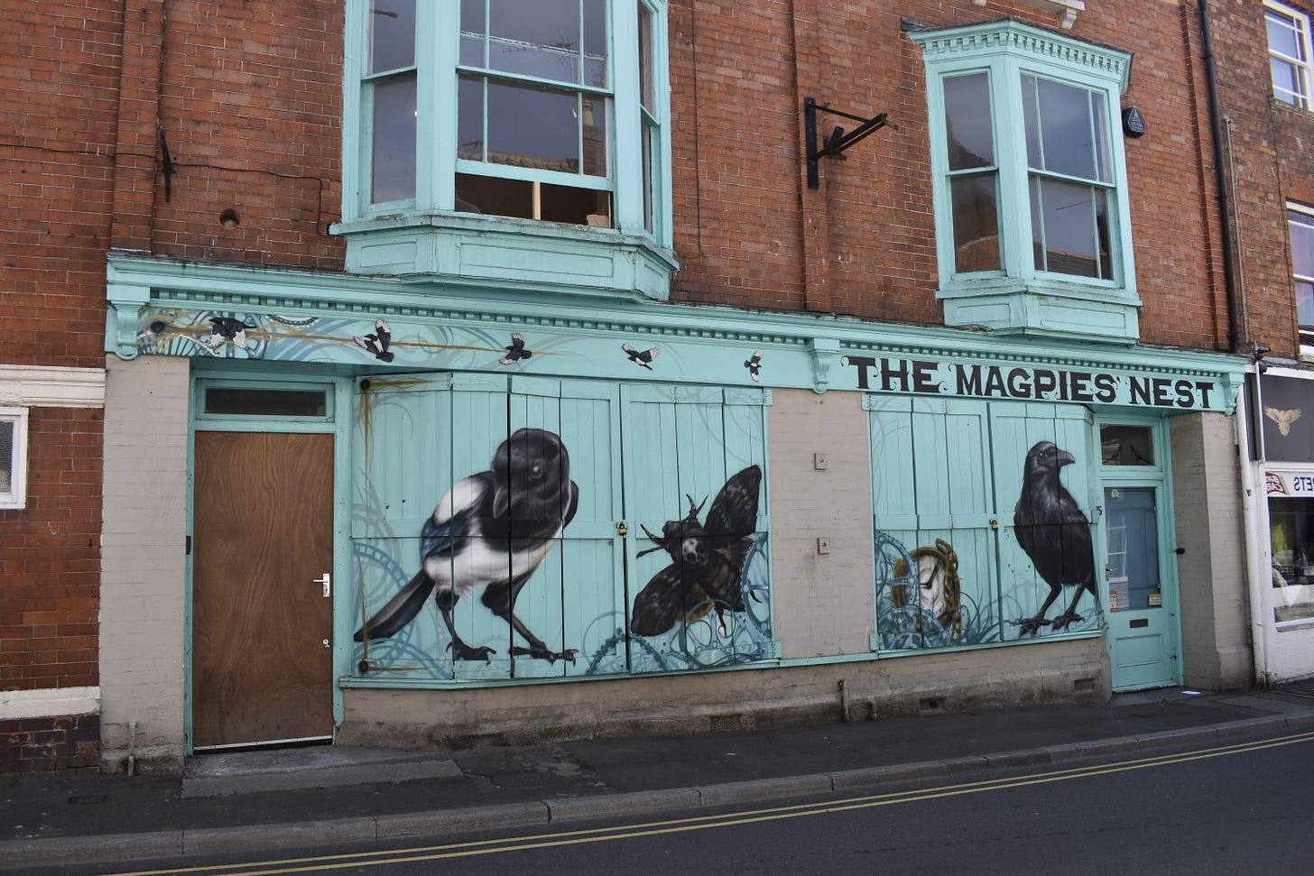 Shuttered shop front with a mural of a blackbird, moth, croww and clock called The Magpies Nest