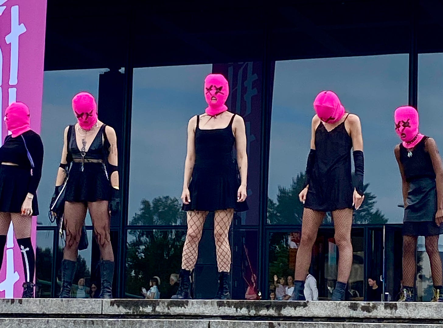 5 mulheres com vestidos preto curto, meia arrastão, todas usando balaclava rosa com X nos olhos.