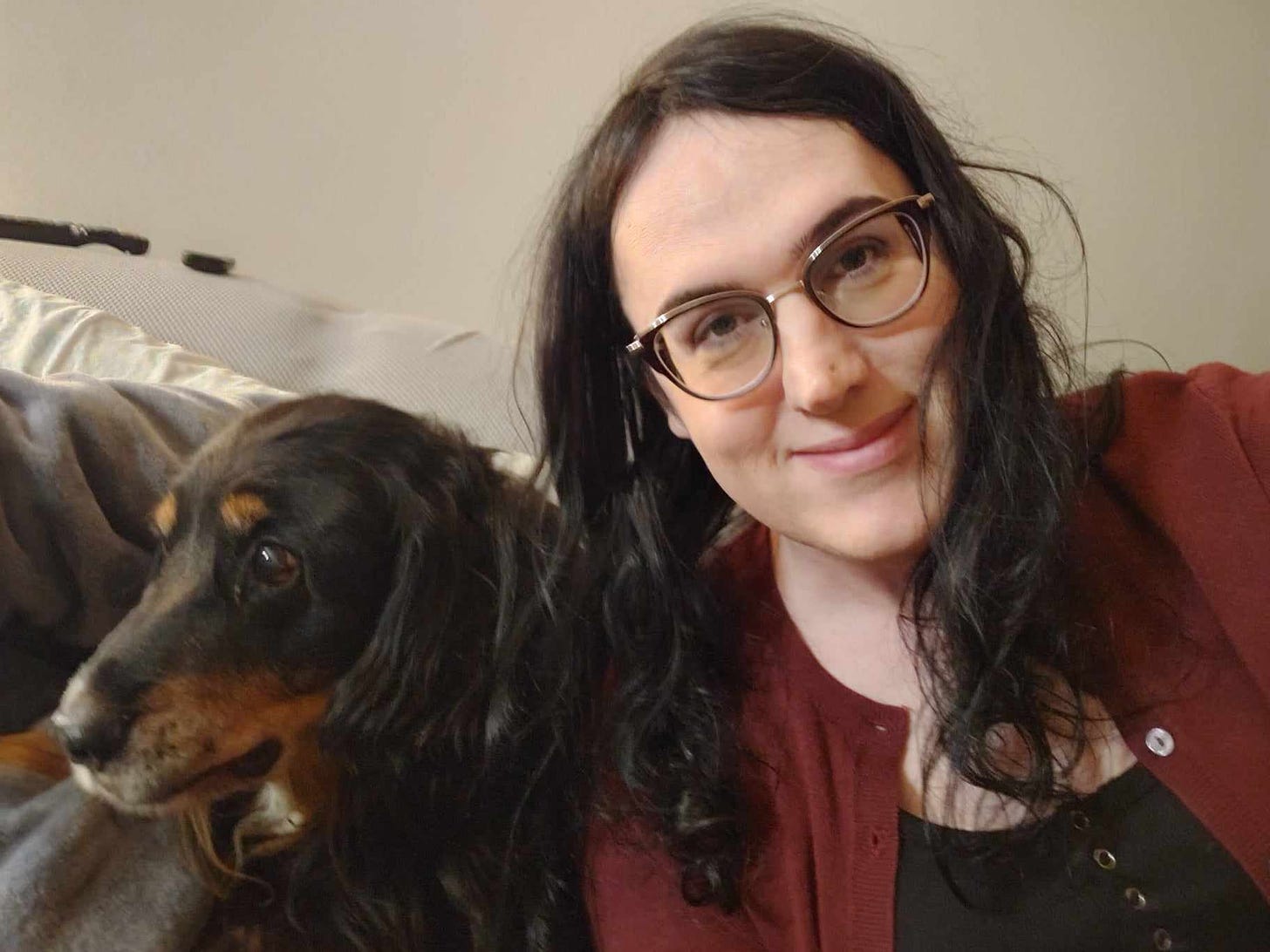 A photo of me, a pale white woman with black horned glasses and wavy black hair, looking exhausted, smiling slightly, next to a black and rust colored dog with wave fur down his ears