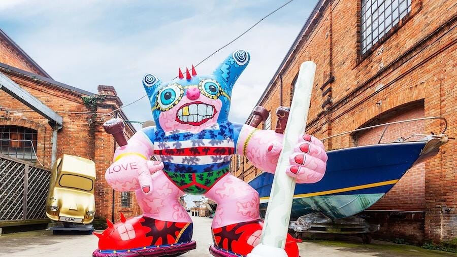 A freaky superhero inflatable sculpture is outside brick buildings. They have a gold tooth, red nails, big spiky red sneakers, tattoos, and brightly colored garments.