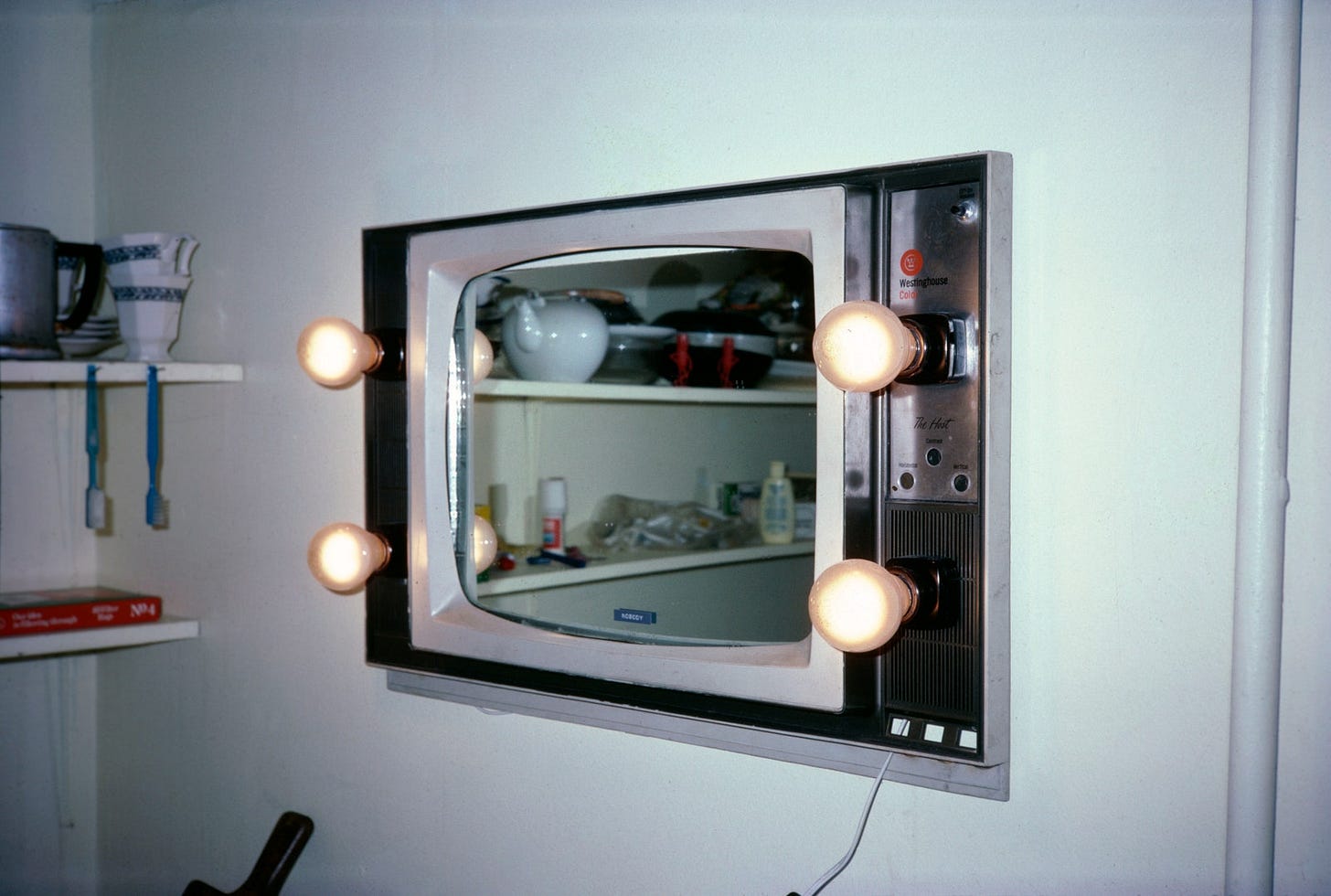 A Westinghouse TV which has been converted to a mirror set into the wall and includes lightbulbs around the perimeter of the screen. The mirror reflects the room and its contents, including teapots, teacups, and two blue toothbrushes hanging from a shelf.