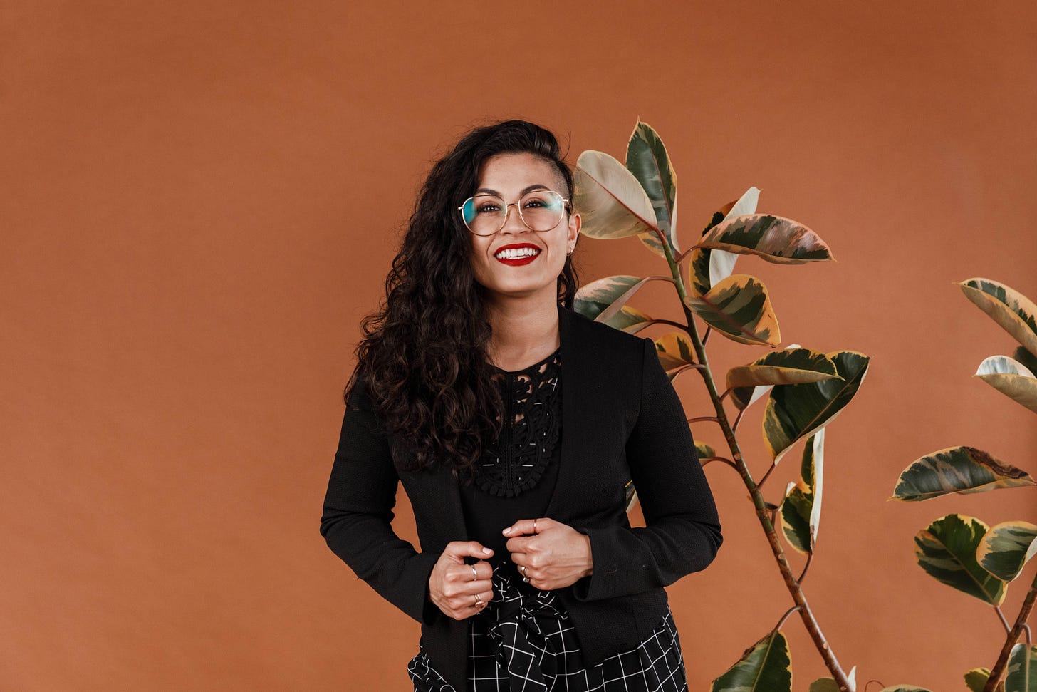 Pam Covarrubias in a black top next to a large plant against a dark orange background