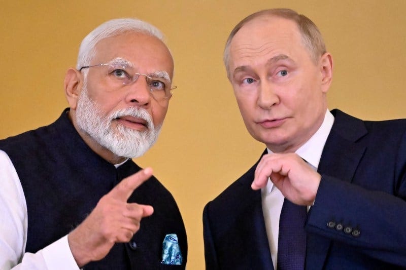 Narendra Modi and Vladimir Putin are seen from the chest upward as they stand near each other in front of a yellow wall. Each man has one hand raised to gesture as they lean close to speak to each other.