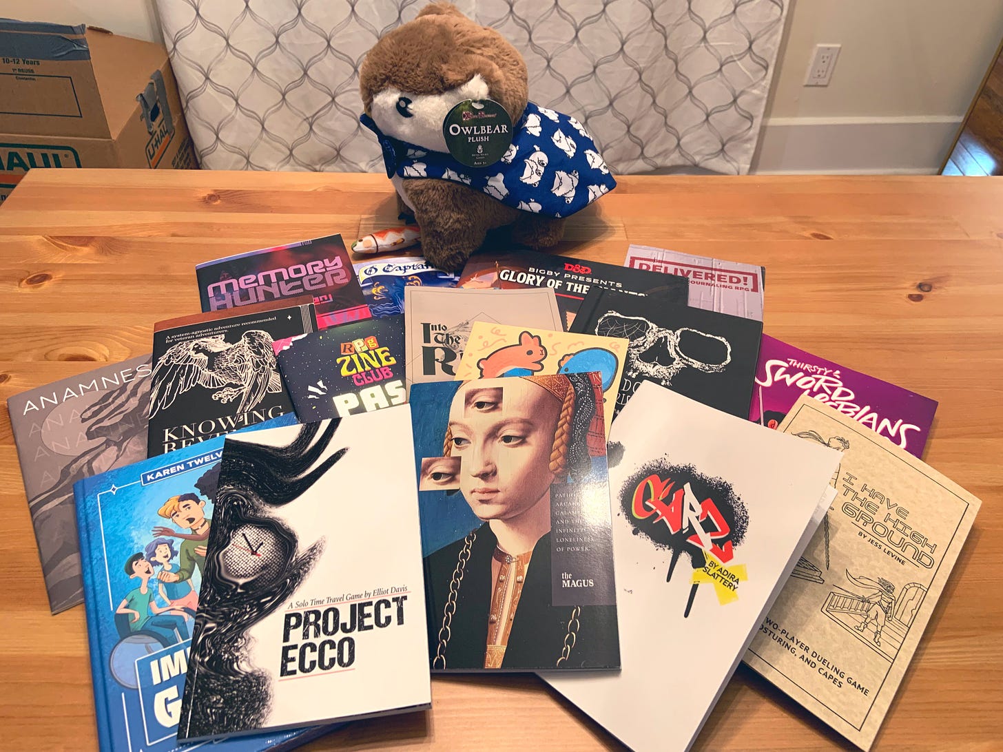 A photo of many TTRPG zines, books, and an owlbear plush toy. The games displayed are: Memory Hunter by Rae Nedjadi, O Captain by Leon Barillaro and Scott Bullock, Bigby Presents: Glory of the Giants by Makenzie De Armas, Dan Dillion, Ben Petrisor, Jason Tondro, and James Wyatt, Delivered by Kari Jo “Kage” Freudigmann, Anamnesis by Sam Leigh, Knowing Revenge of the Grasping Gods by Kyle Tam, Zine Club Passport by Plus One Exp, Into the Riverlands by Josh Hittie, Down We Go by Markus Linderum, Thirsty Sword Lesbians by April Kit Walsh, Improv for Gamers by Karen Twelves, Project Ecco by Elliot Davis, The Magus by momatoes, CARZ by Adira Slattery, and I Have the High Ground by Jess Levine.