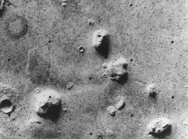 A grainy black-and-white image of the Martian surface, taken from above, showing several partially shadowed hills. The pattern of shadows on one hill resembles a human face.