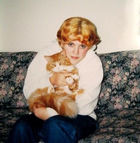 a young person with short curly orange hair sits on a couch holding an orange tabby cat