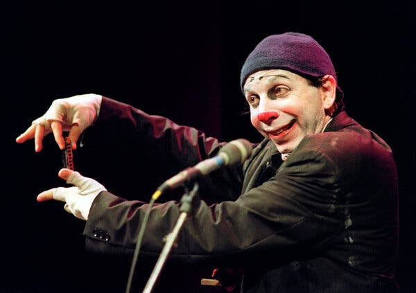 A smiling clown with a white face, a red nose and a purple cap stands at a microphone holding a harmonica in both hands.