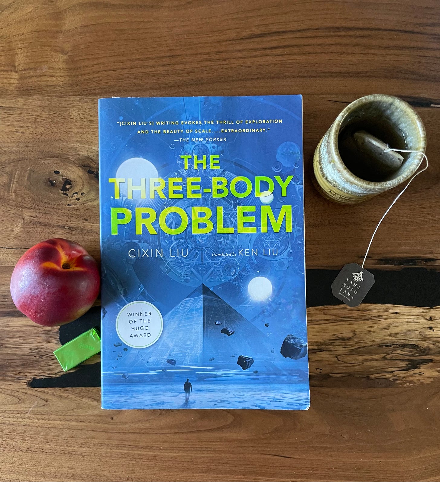 A picture of the book The Three Body Problem, staged on a wooden table with a mug of tea, a nectarine, and a square of Meiji matcha chocolate