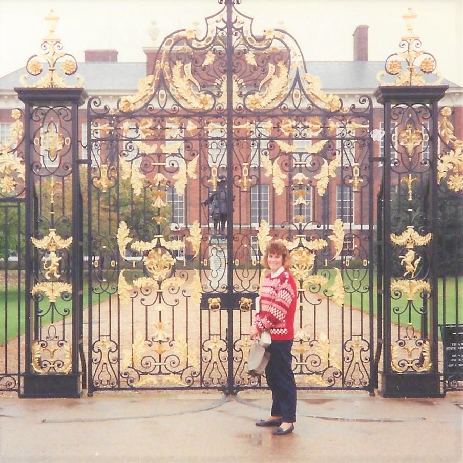 20-something woman stands outside the very ornate gates of Kensington Palace