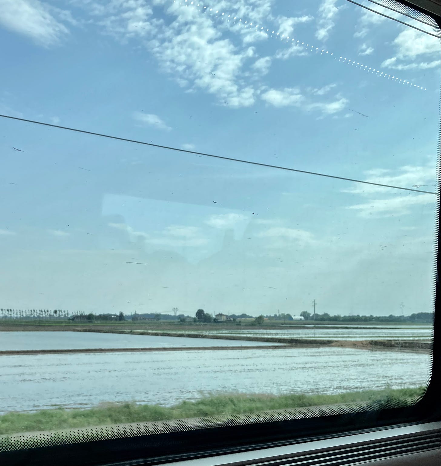 Da janela do trem, duas plantações inundadas de água. É possível ver o céu e a janela do trem.
