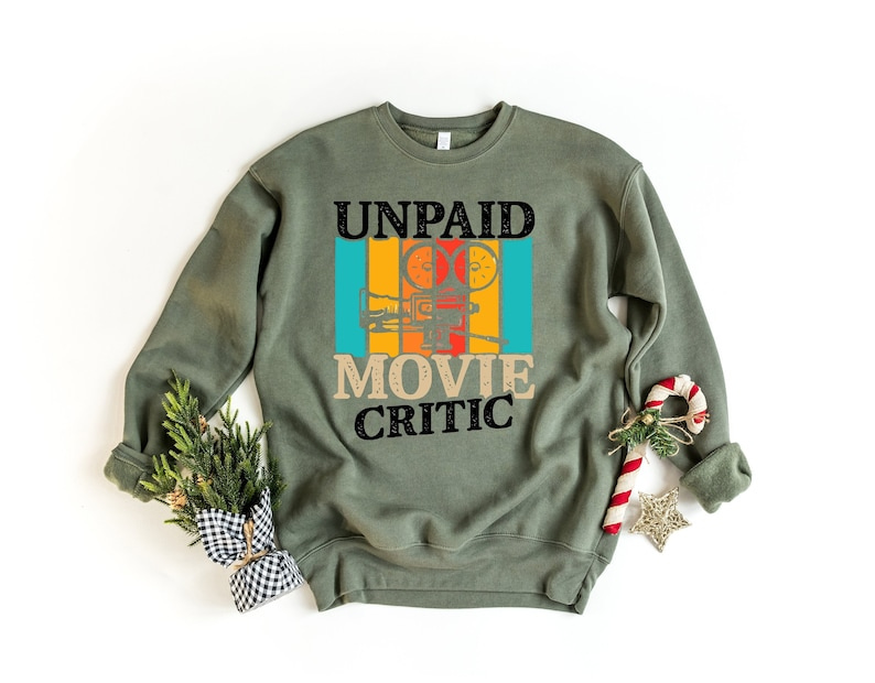 May include: A green sweatshirt with the text 'UNPAID MOVIE CRITIC' in black and white. The text is surrounded by a colorful retro-style design of a film projector.