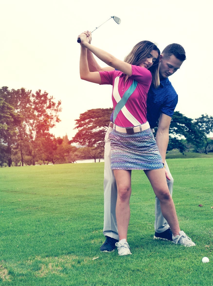 Woman i purple blouse raising golf club on backstroke with man touching her leg, teachng her to swing