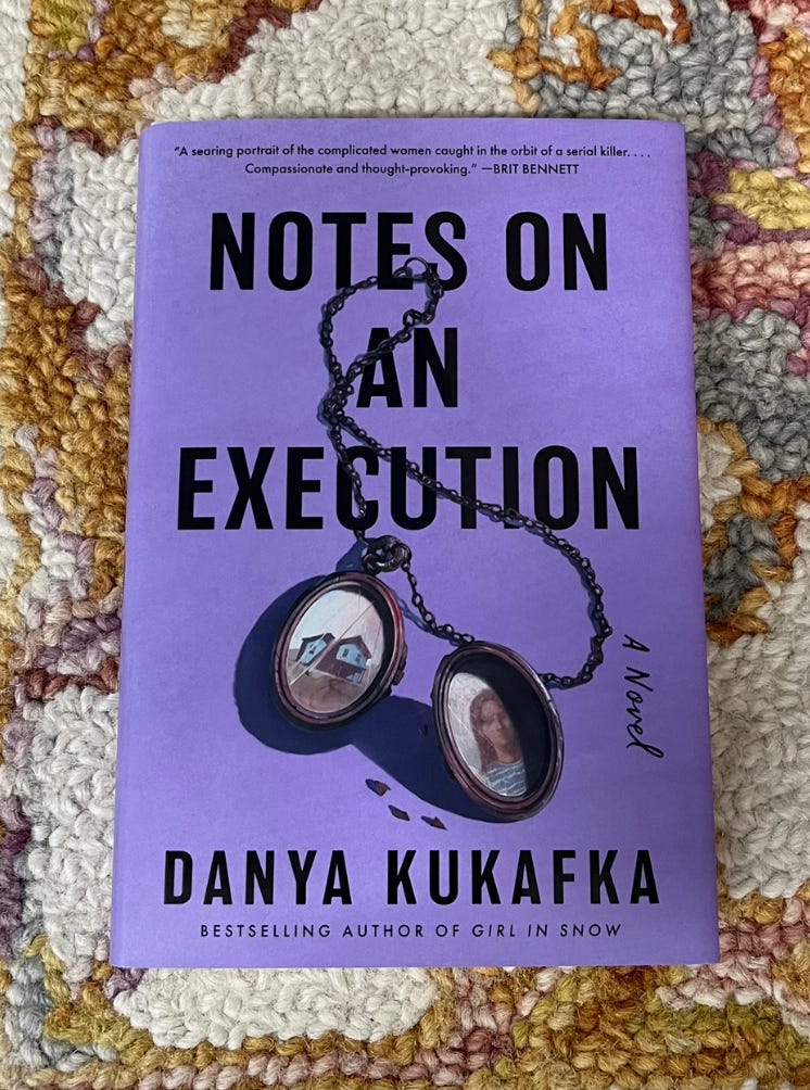 Rich purple cover with NOTES ON AN EXECUTION title, drawing of locket, and Danya Kukafka's name. Book is laying on a rug.