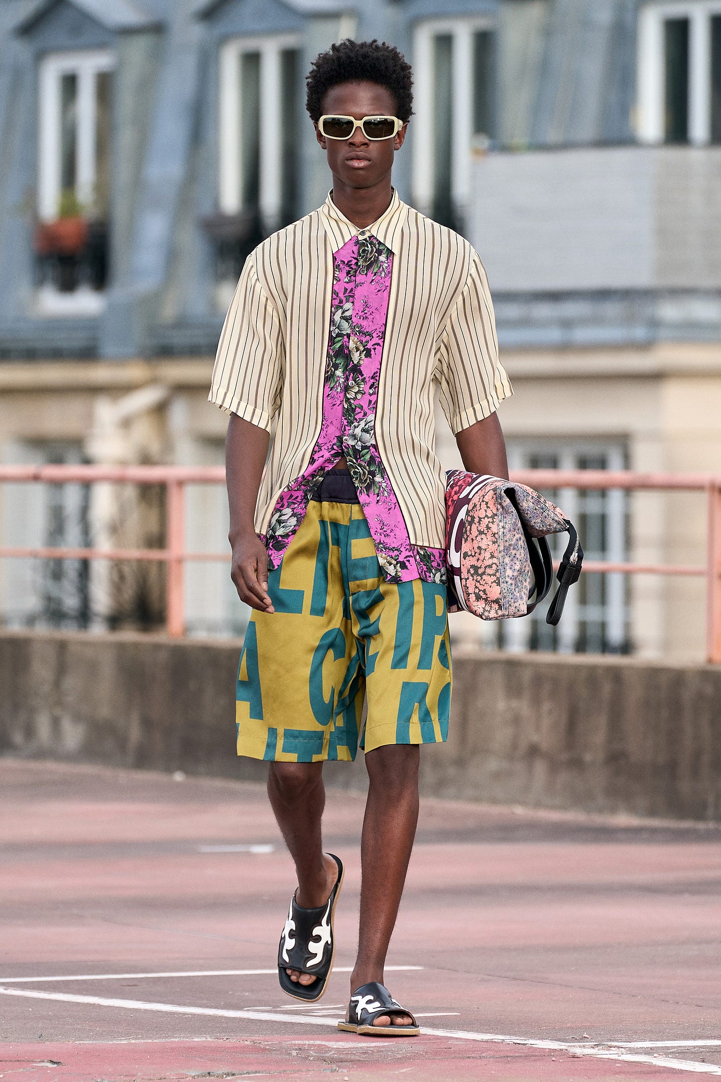 Black male model on the catwalk wearing a buttoned-up short-sleeved cream striped shirt with contrasting pink floral placket and bold print shorts.